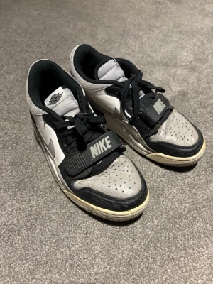 Nike Air Jordan grå/svarta sneakers - Nike Air Jordan sneakers i grått, svart och vitt med klassisk Nike-rem över vristen och Jordan-logga på hälen. Ovandelen är i läder med perforerade detaljer och svart snörning. Snygg kontrast mellan färgerna och ikoniska detaljer som ger en sportig vibe. Ett par gamla skor som jag nu säljer 