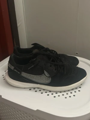 Svarta sneakers med grå detalj futsal tiempo - Snygga svarta sneakers med grått swoosh-märke på sidan och vita sulor. Skorna har snörning och en låg profil som passar till många olika stilar. Materialet ser ut att vara en mix av textil och syntet. Perfekta för dig som gillar en sportig look.