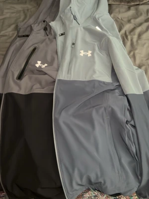 Bara gråa kvar - Snygg grå vindjacka från Under Armour med huva och dragkedja framtill. Jackan har en sportig look med logga på bröstet och praktiska fickor. Perfekt för blåsiga dagar och outdoor-aktiviteter.
