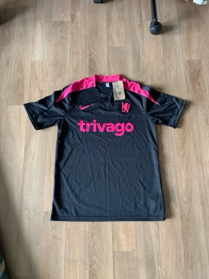 Snygg Chelsea fc tröja  - Snygg svart fotbollströja från Nike med rosa detaljer och tryck. Tröjan har korta ärmar, rosa axelpartier och logga samt trivago-tryck på bröstet. Perfekt för träning eller match och har en sportig, modern look.