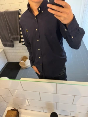 Ralph Lauren skjorta - Säljer en mörkblå Ralph Lauren skjorta med klassisk button-down krage och vita knappar. Skjortan har lång ärm och en broderad gul logga på bröstet. Perfekt för dig som gillar stilrena plagg med enkel design.