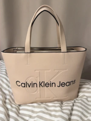 Beige handväska Calvin Klein Jeans - Stilren beige handväska från Calvin Klein Jeans med präglad CK-logga och svart text på framsidan. Väskan har två handtag, dragkedja upptill och är gjord i slätt konstläder. Perfekt för dig som gillar minimalistisk design och vill ha plats för allt du behöver.