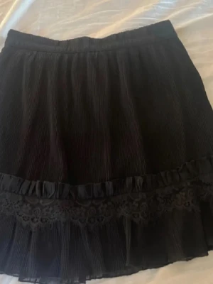 Svart kortkjol med spets och volang - Säljer en svart kortkjol med plisserad struktur och dekorativ spetsdetalj samt volang med spets längst ner. Kjolen har en elastisk midja och ger en söt och trendig look. Perfekt att matcha med en enkel topp eller stickad tröja. Super söt nu till höst🤎