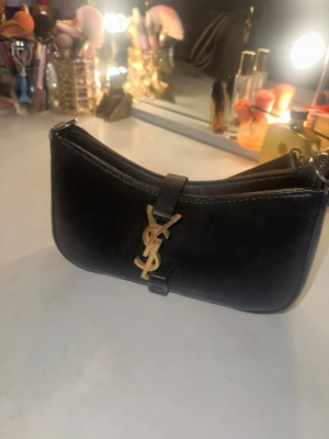 Svart axelväska med guldiga detaljer - Snygg svart handväska i skinn med axelrem och ikonisk guldig YSL-logga framtill. Väskan har en stilren, avlång form och stängs med ett band över loggan. Perfekt accessoar för att lyfta vilken outfit som helst. 