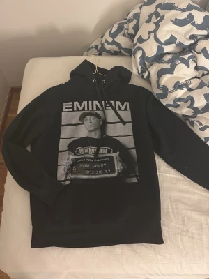 Svart Eminem hoodie med tryck - Svart hoodie med stor vit Eminem- och Slim Shady-tryck på framsidan. Motivet föreställer Eminem i mugshot-stil med texten 'RUNYON AVE' och 'Detroit Police Department'. Klassisk huva med dragsko och långa ärmar. Perfekt för dig som gillar streetwear och hiphop. De finns ingen lapp om storlek men den passar nog S/M då jag bär kläder i storlek M och den passa mig ganska på gränsen💕