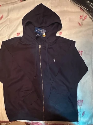 Mörkblå hoodie zipper från Polo Ralph Lauren oanvänd helt ny - Mörkblå hoodie med dragkedja från Polo Ralph Lauren. Klassisk design med huva, fickor framtill och den ikoniska vita logotypen broderad på bröstet. Tillverkad i mjukt bomullsmaterial som är skönt att bära. Perfekt för en avslappnad och stilren look.