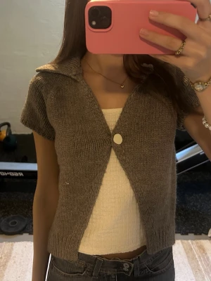 Kort stickad brun kofta med krage - Kortärmad stickad kofta i brun nyans med krage och en stor knapp framtill. Modellen är croppad och har en mjuk, mysig känsla som passar perfekt till jeans eller kjol. Materialet ser ut att vara ullblandning eller liknande för en skön och avslappnad look.