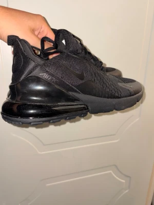 Svarta Nike Air Max 270 sneakers - Säljer ett par helsvarta Nike Air Max 270 sneakers med synlig Air-enhet i hälen och mesh-ovandel. Skorna har en sportig look med diskret Swoosh-logga på sidan och slitstark gummisula. Perfekta för dig som gillar streetwear och bekväma skor.