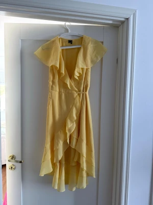 Gul omlottklänning med volang - Snygg gul omlottklänning med volangdetaljer och knytband i midjan. Klänningen har v-ringning, ärmlös design och faller i en längre modell med asymmetrisk nederkant. Perfekt för sommarens alla tillfällen.