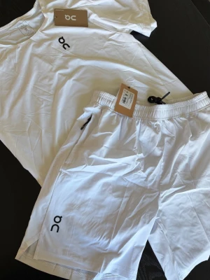Vitt träningsset från On - Säljer ett fräscht vitt träningsset från On med t-shirt och shorts. Båda plaggen har minimalistisk design med On-logga tryckt framtill. Shortsen har elastisk midja och dragkedjeficka. Perfekt för gym eller löpning.