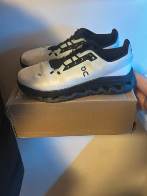 Vita On running - Snygga vita On Cloudmonster sneakers med svart sula och svarta detaljer. Skorna har en chunky, vågig sula och ventilerande meshmaterial på ovandelen. Loggan 'On' syns tydligt på sidan och tån. Perfekta för dig som gillar sportig och modern stil.