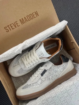 Glittriga sneakers från Steve Madden - Snygga sneakers från Steve Madden i vitt och silver med glittriga detaljer och beige insida. Skorna har vita snören, en brun sula och en mix av mocka och textilmaterial. Perfekta för dig som vill sticka ut med stilrena men ändå unika skor.