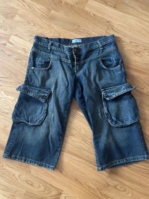 Lågmidjade cargoshorts/jorts i denim - Säljer ett par lågmidjade blå cargojorts i denim med stora fickor på sidorna! Ca 41cm rakt över midjan 💕