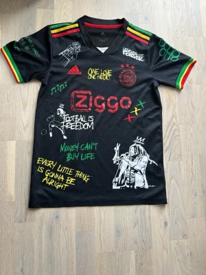 Svart Ajax x Bob Marley tröja - Unik svart Ajax-tröja från Adidas med Bob Marley-tema. Flera färgglada tryck och citat, illustrationer av Bob Marley och reggaeinspirerade detaljer i rött, gult och grönt på axlar och ärmslut. Perfekt för dig som gillar fotboll och musik.