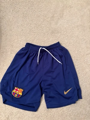 Blå FC Barcelona shorts från Nike - Snygga marinblå shorts från Nike med FC Barcelona-logga på ena benet och gul Nike-swoosh på det andra. Shortsen har resår i midjan och vit snörning för bästa passform. Perfekta för fotboll eller träning. Storlek S-M