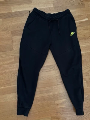 Svarta Nike mjukisbyxor med logga - Säljer ett par svarta mjukisbyxor från Nike med gul logga på vänster ben. Byxorna har dragsko i midjan, muddar vid bensluten och två sidofickor. Perfekta för chill dagar eller träning. Sköna och stilrena med modern passform.