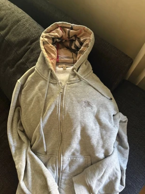 Grå hoodie med dragkedja och mönstrad huva - Snygg grå hoodie med dragkedja och klassisk känguruficka. Huvan har ett rutigt innerfoder i beige, svart och rött. Broderad logga på bröstet och justerbara snören i huvan. Perfekt för en chill och avslappnad stil. Står L men passar både större M och L