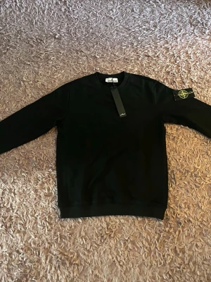 Svart sweatshirt från Stone Island - Säljer en svart sweatshirt från Stone Island med den klassiska loggan på vänster ärm. Tröjan har rund halsringning och ribbade muddar vid ärmslut och nederkant. Perfekt för dig som gillar streetwear och vill ha en stilren look.