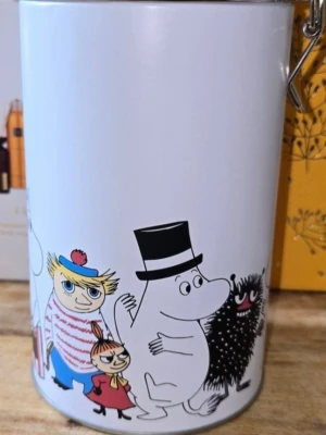 Mumin burk med färgglada figurer - Vit plåtburk med motiv av Mumin och vänner runt om. Burken har ett tättslutande lock med metallspänne och färgglada illustrationer av klassiska Mumin-karaktärer. Perfekt för förvaring av kaffe, te eller småsaker.