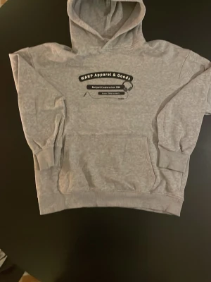 Grå hoodie från WARP Apparel & Goods - Snygg grå hoodie med tryck på bröstet från WARP Apparel & Goods. Texten på framsidan lyder 'Backyard Creators since 1996' och har ett motiv av hörlurar. Hoodien har huva och magficka, samt ribbade muddar vid ärmar och nederkant.