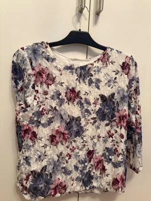 blommig topp med trekvartsärm - Söt blus i vit spets med blommigt mönster i blått, lila och rosa toner. Blusen har trekvartsärm och rund halsringning. Perfekt för dig som gillar romantisk och feminin stil.