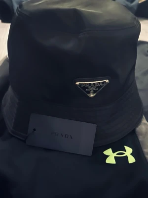 Prada bucket  - Snygg svart bucket hat från Prada med klassisk trekantig metallogo framtill. Hatten är tillverkad i ett glansigt, slitstarkt material och har diskreta sömmar runt brättet. Perfekt accessoar för en trendig och stilren look.