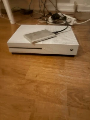 Vit Xbox One S spelkonsol - Säljer en vit Xbox One S spelkonsol med stilren design och matta ytor. Konsolen har en diskret svart detalj och är tillverkad i plast. En extern hårddisk är ansluten via USB-porten på ovansidan. Perfekt för gaming och streaming.