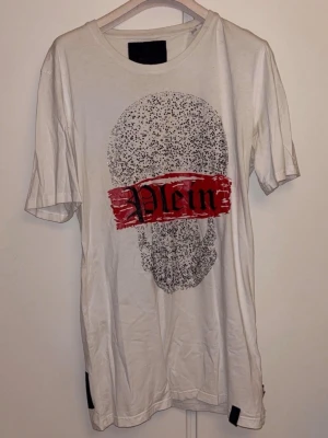 Philip plein t-shirt med döskalle av kristaller  - Säljer en vit Philip plein med ett stort tryck av en  döskalle på framsidan och en röd text.  'Plein' över. T-shirten har rund hals och korta ärmar. Perfekt för dig som gillar  streetstyle.