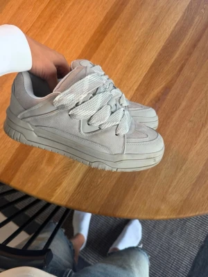 Gråa bershka skor - Stilrena chunky sneakers i helvitt med breda, platta snören och kraftig sula. Skorna har en modern siluett med paneler i textil och syntetmaterial. Perfekta för dig som gillar en clean och trendig look.