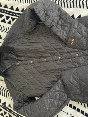 Barbour jacka - Svart quiltad jacka från Barbour med klassisk krage i manchestertyg och tryckknappar framtill. Jackan har Barbour-logga broderad på bröstet och innerficka med dragkedja. 