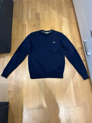 Mörkblå långärmad tröja Lacoste - Snygg mörkblå långärmad tröja från Lacoste med klassisk rund halsringning och den ikoniska krokodilloggan på bröstet. Tröjan har en stilren och enkel design, perfekt att matcha med jeans eller kjol.