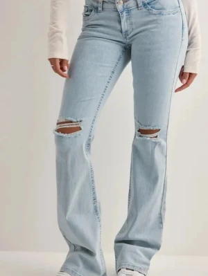 Ljusblå bootcut jeans med slitningar - Säljer ett par ljusblå bootcut jeans med slitna hål över knäna och broderade detaljer på bakfickorna. Jeansen har hög midja och klassisk femficksdesign. Tillverkade i denim med en avslappnad passform och trendig look.