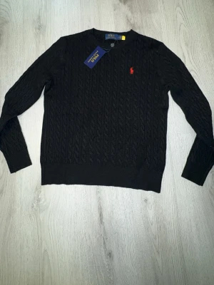 Svart kabelstickad tröja Polo Ralph Lauren - Kabelstickad svart tröja från Polo Ralph Lauren med rund halsringning och den klassiska röda logotypen broderad på bröstet. Tröjan har långa ärmar och ribbade muddar. Perfekt för dig som gillar stilrena och tidlösa plagg.