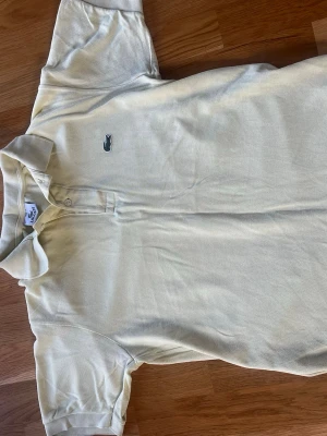 Ljusgul piké från Lacoste - Säljer en klassisk ljusgul pikétröja från Lacoste med den ikoniska krokodilloggan på bröstet. Tröjan har korta ärmar, krage och knappar vid halsen. Tillverkad i mjuk bomull som känns skön mot huden. Perfekt för en avslappnad och stilren look.