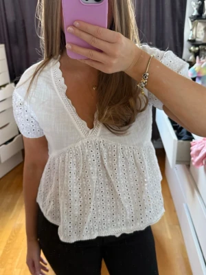 Vit blus med spets och broderi - Söt vit blus med v-ringning, korta puffärmar och broderade detaljer. Blusen har spetskant längs ringningen och är tillverkad i ett lätt bomullstyg med hålbroderi. Perfekt för en fräsch och somrig look.