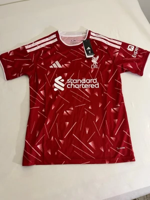 Liverpool röd matchtröja Adidas - Snygg röd Liverpool matchtröja från Adidas med vitt geometriskt mönster och vita detaljer på axlarna. Tröjan har klubbmärke och sponsortryck framtill samt korta ärmar. Perfekt för fotbollsträning eller att bära till vardags.