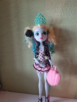 Monster High maskerad docka - Cool Monster High-inspirerad maskerad docka med blått och blont lockigt hår, turkos tiara och svart blomaccessoar. Hon har en vit klänning med rosa och svarta detaljer, samt en rosa väska formad som ett snäckskal. Perfekt för cosplay eller maskerad.