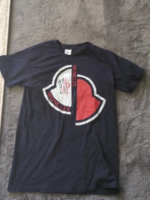 Svart Moncler t-shirt med stor logga - Snygg svart t-shirt från Moncler med stor, delad logga i vitt och rött på bröstet. T-shirten har klassisk passform och rund hals. Perfekt för dig som gillar streetwear och vill sticka ut med ett tydligt märke.