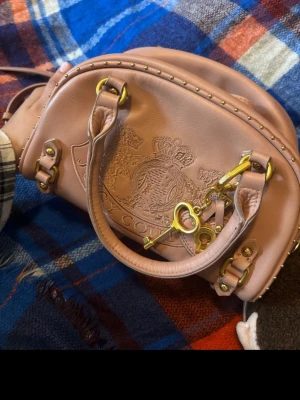 Rosa handväska från Juicy Couture - Säljer en snygg rosa handväska från Juicy Couture med guldfärgade detaljer och nitar. Väskan har broderad logga framtill, två handtag och en dekorativ nyckel. Perfekt för dig som gillar Y2K-stil och vill sticka ut.