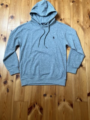 Grå hoodie från Polo Ralph Lauren - Klassisk grå hoodie från Polo Ralph Lauren med svart broderad logga på bröstet. Hoodien har huva med dragsko, ribbade muddar och en mjuk bomullsblandning. Perfekt för en avslappnad och stilren look.
