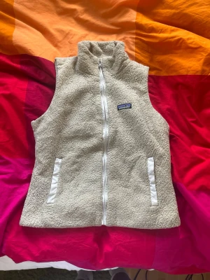 Beige teddyväst från Patagonia - Mysig beige teddyväst från Patagonia med hög krage och dragkedja framtill. Västen har två fickor med vita detaljer och är gjord i mjukt, fluffigt material som ger en trendig look. Perfekt att slänga över en hoodie eller tröja för extra värme.
