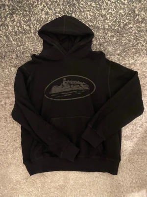 Svart hoodie med tryck av byggnad - Säljer en svart hoodie med huva och ett stort tryck av en byggnad i vitt på bröstet. Hoodien har klassisk känguruficka framtill och ribbade muddar vid ärmar och nederkant. Perfekt för dig som gillar stilrena och bekväma plagg.