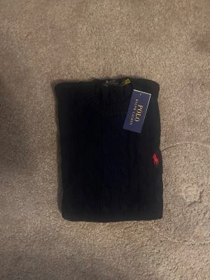 Mörkblå kabelstickad tröja Polo Ralph Lauren - Snygg mörkblå kabelstickad tröja från Polo Ralph Lauren med klassisk röd logga broderad på bröstet. Tröjan har rund halsringning och ribbade muddar vid ärmslut och nederkant. Perfekt för dig som gillar stilrena och tidlösa plagg.