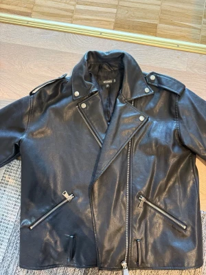 Svart biker skinnjacka från Missguided - Snygg svart biker skinnjacka från Missguided med silvriga dragkedjor och knappar. Klassisk modell med bred krage, axelklaffar och flera fickor med dragkedja. Perfekt för en edgy och trendig look.