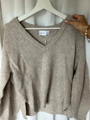 Beige stickad tröja från VILA - Mysig beige stickad tröja från VILA med v-ringning och avslappnad passform. Tröjan har långa ärmar och ribbade muddar vid hals, ärmslut och nederkant. Perfekt för lager på lager och enkel att matcha med jeans eller kjol.