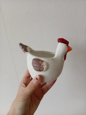 Dekorativ höna i keramik - Rolig och charmig liten höna i keramik, perfekt som dekoration eller liten kruka. Hönan är vit med bruna vingar, röd kam och orange näbb. Passar fint i köket eller på påskbordet för en extra touch av humor och personlighet. Finns nå märken