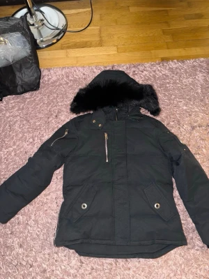Svart dunjacka från moose knuckles - Snygg svart dunjacka med huva och svart fuskpäls. Jackan har dragkedja framtill, två fickor med lock och knappar samt en bröstficka med dragkedja. Detalj i metall på ena ärmen. Perfekt för kalla vinterdagar.