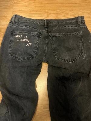 Svarta jeans med broderad text - Säljer ett par svarta jeans från Gina i storlek 36. Jeansen har en broderad text på bakfickan där det står 'WHAT U LOOKIN AT' och ett slitet parti på ena benet. Klassisk femficksmodell och raka ben.