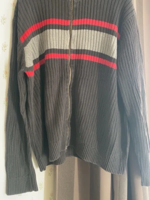 Mörkgrå stickad ziptröja Tommy Hilfiger - Mörkgrå stickad ziptröja från Tommy Hilfiger med ribbad struktur och färgblock i rött, vitt och svart över bröstet. Liten logga på ärmen. Tröjan har långa ärmar och dragkedja framtill. Perfekt för lager-på-lager-stil.