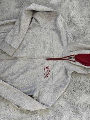 Hugo Boss zip hoodie - Snygg grå zip-hoodie från BOSS med röd insida på huvan och röda snören. Dragkedja framtill, fickor på sidorna och BOSS-logga broderad på bröstet. Perfekt för en chill och stilren look.